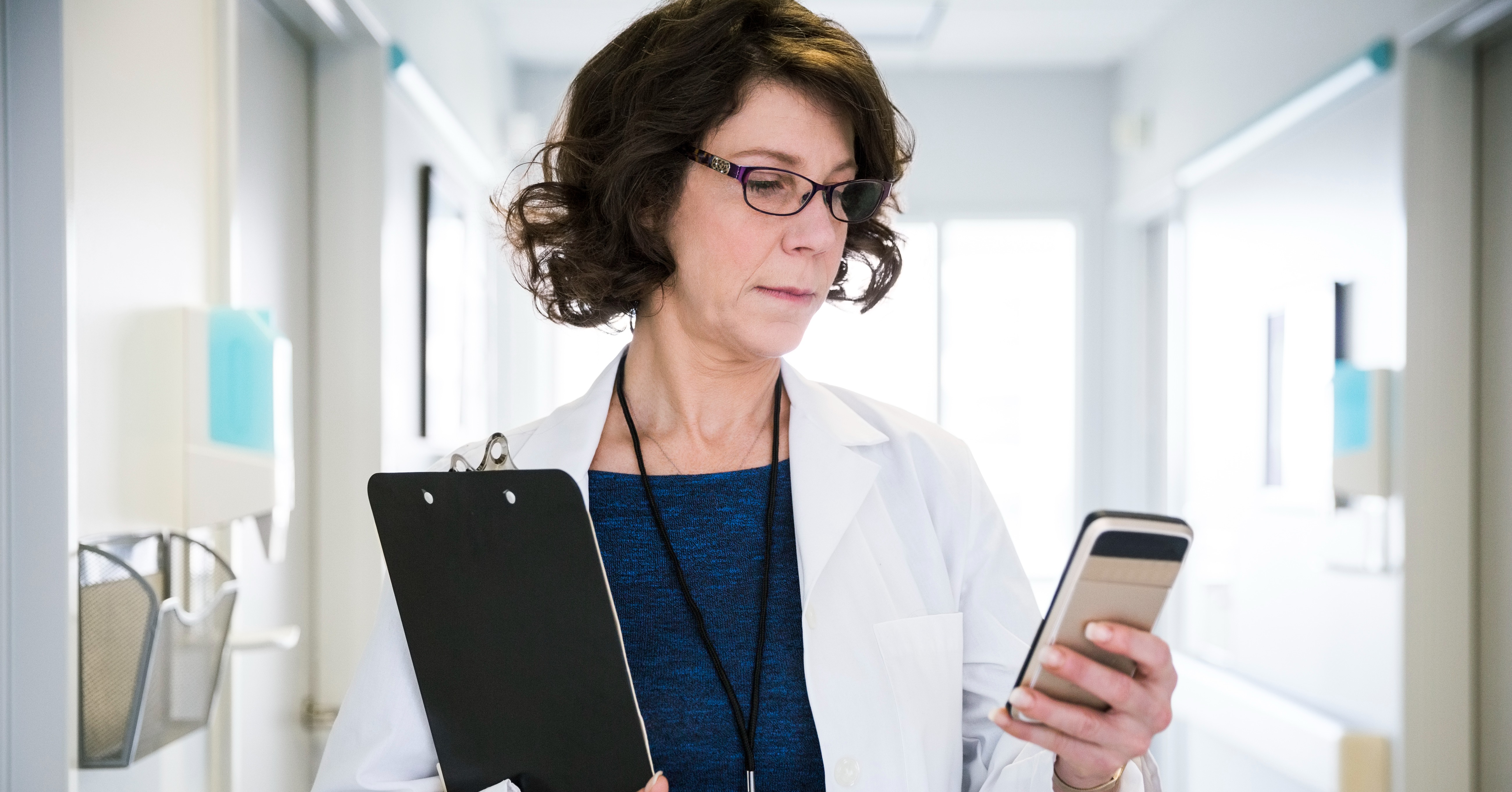 Physician looks down at her smartphone while holding a clipboard